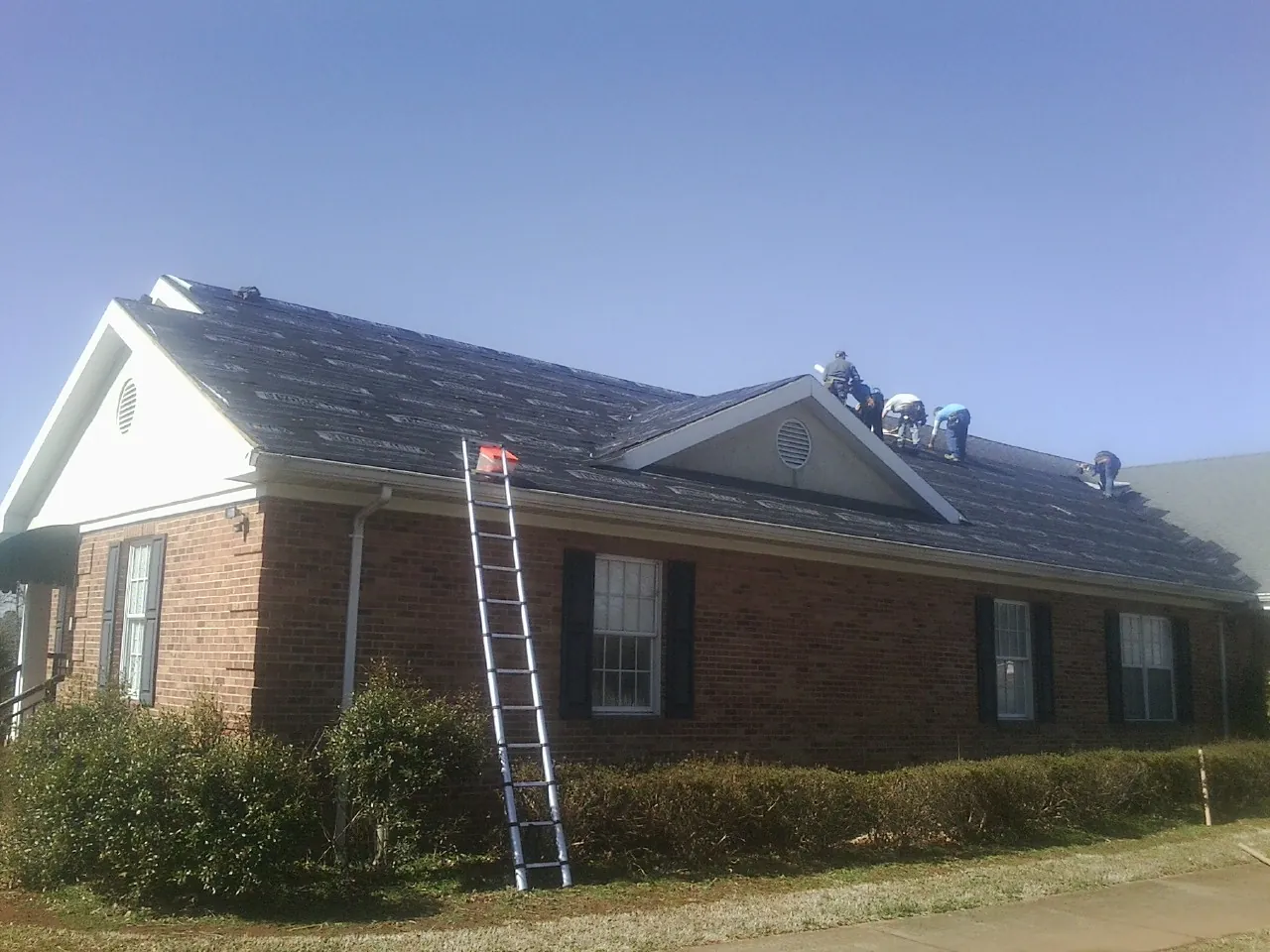 Crew tearing shingles off a brick ranch roof with a ladder set — Emergency Roof Repair work by our crew in Gould