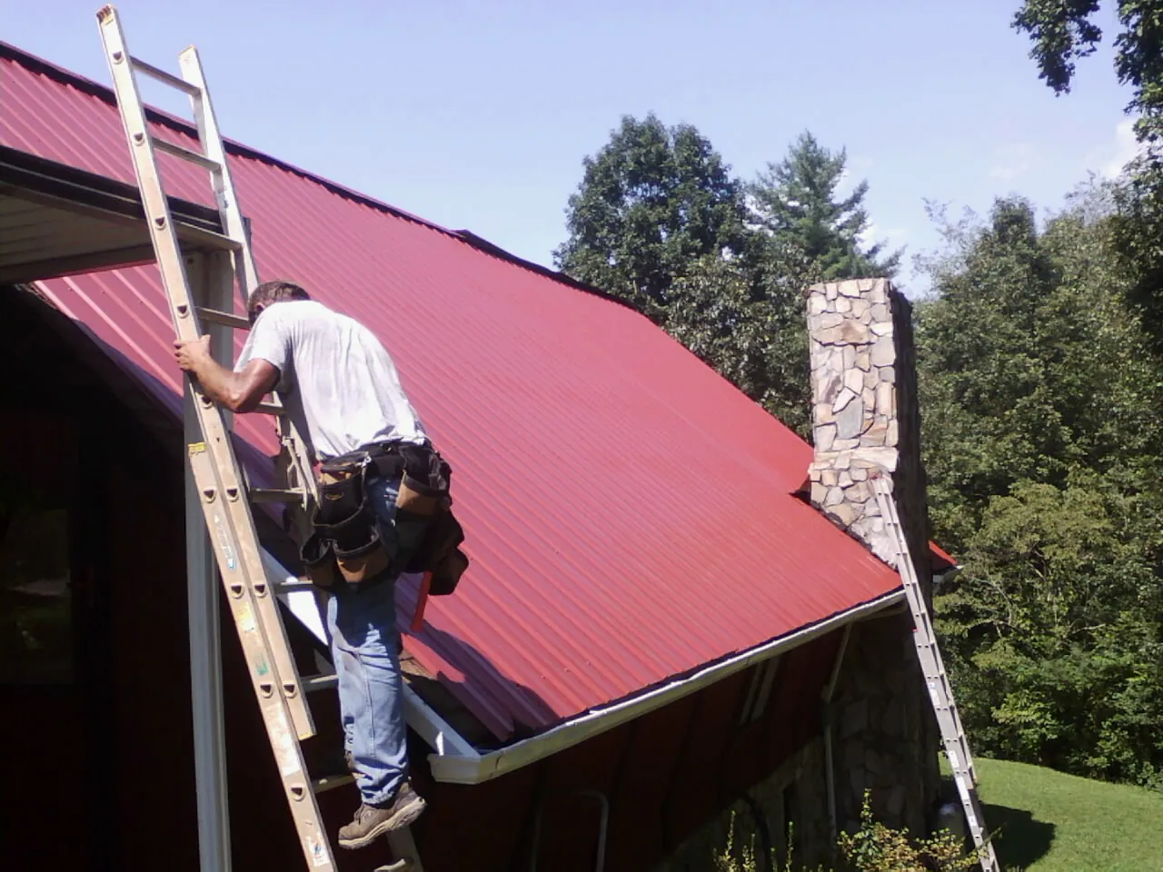 Roofer on a ladder beside a red metal roof and stone chimney — Soffit & Fascia Repair by Roofing Brothers in Gould